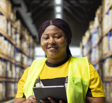 African american staff in workwear uses device to manage product handling, controlling inventory in warehouse with storage racks. Parcels organized for local delivery, maintaining high standards.