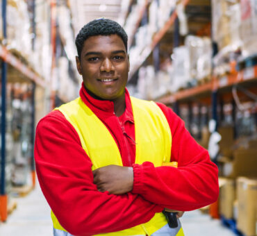 Confident warehouse worker smiling with arms crossed, holding a barcode scanner in a logistics center, surrounded by shelves